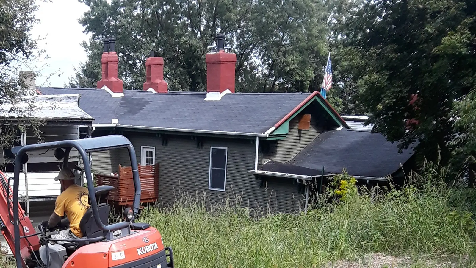 Operator working a mini excavator in an overgrown lot beside the house that is coming down