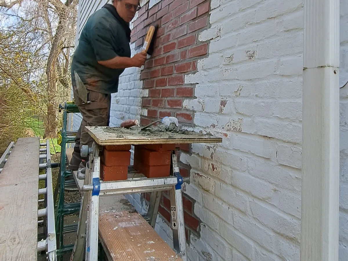 Mason on a work plank laying new brick into an opening in a painted brick wall