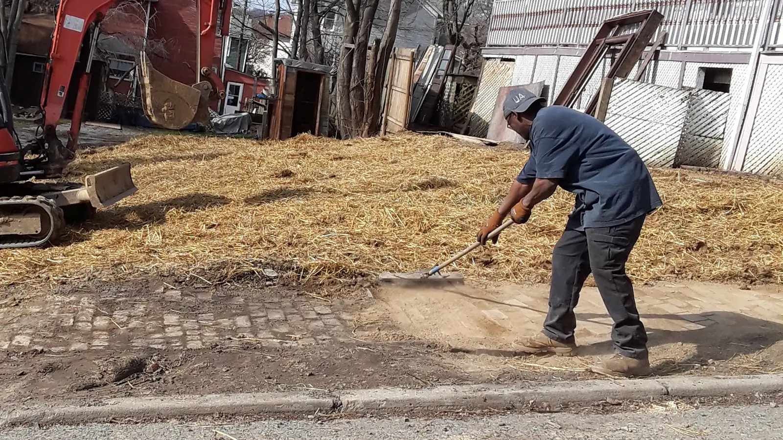 Mini excavator parked on a cleared demolition lot while a worker rakes straw over the fresh grade in the Pittsburgh area